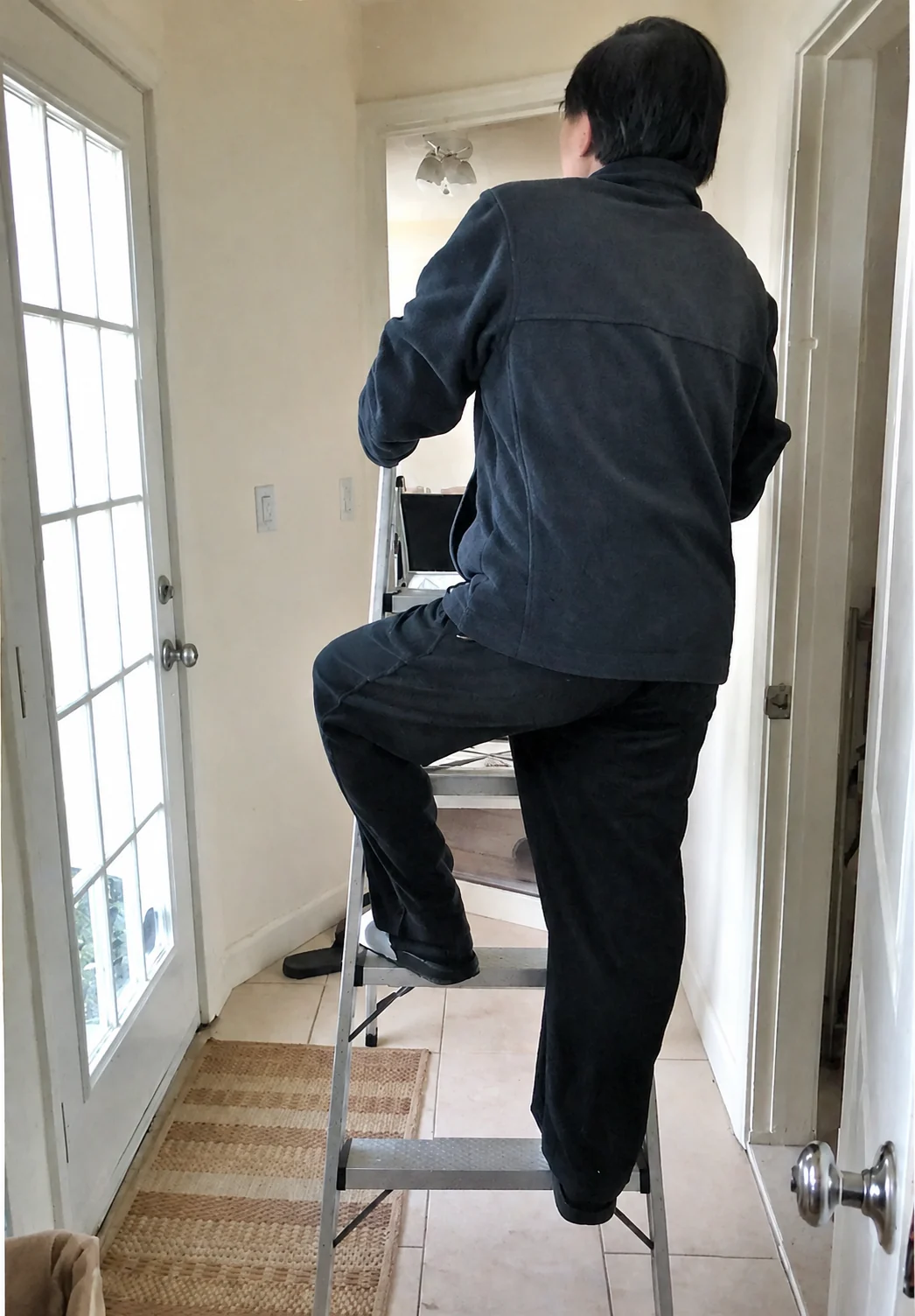 A worker stands on a ladder in a narrow room while painting a wall.